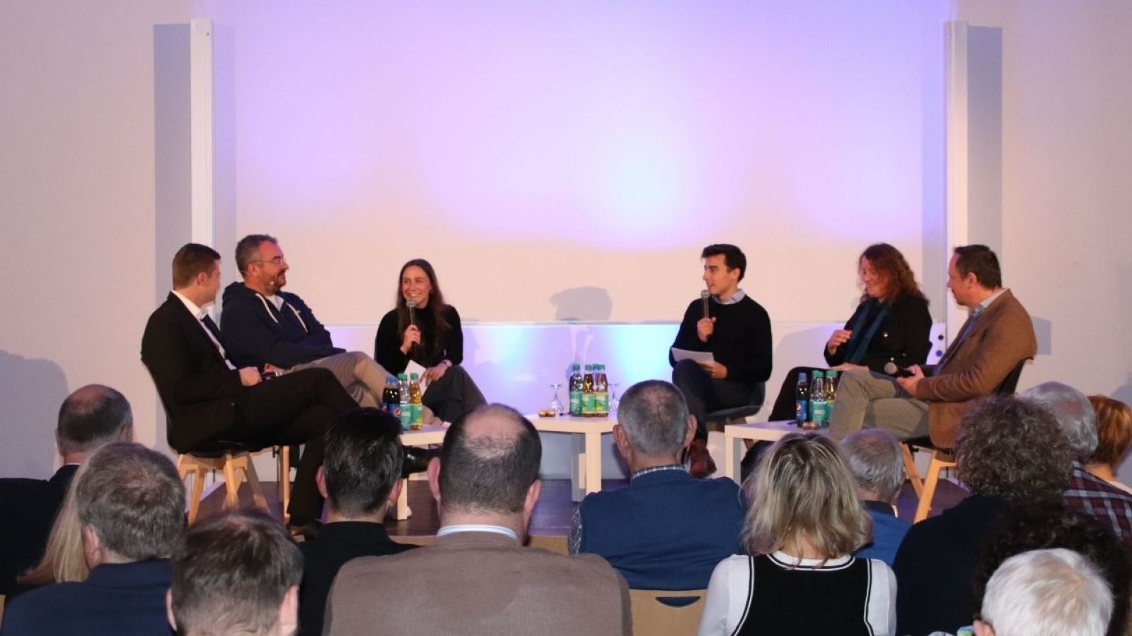 Podiumsdiskussion mit sechs Teilnehmern vor Publikum bei einer Konferenz, ausgeleuchtet mit blauem Licht.