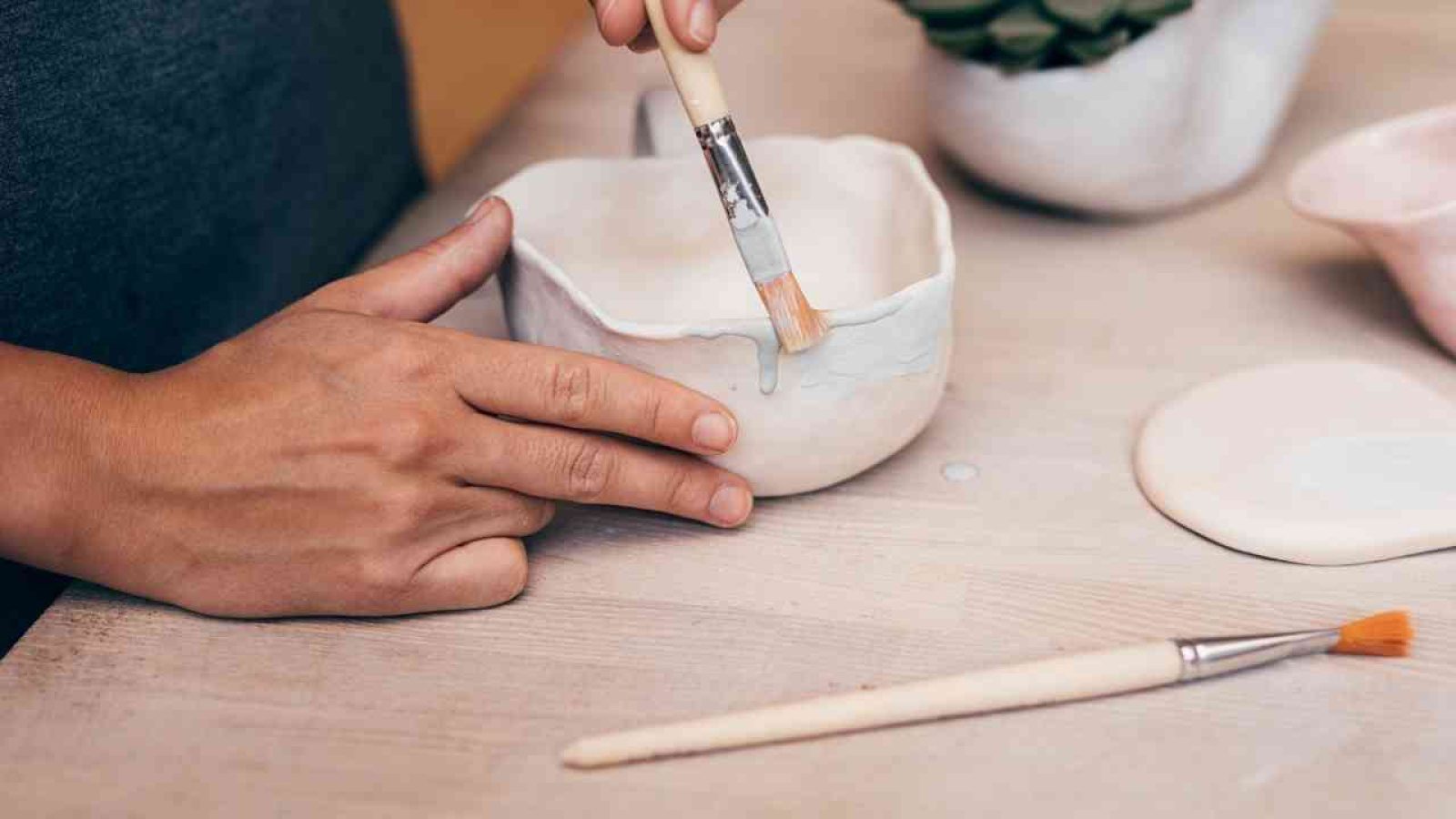 Person bemalt handgefertigte Keramikschüssel mit Pinsel auf Holztisch. Künstlerische Handarbeit und Keramikgestaltung.