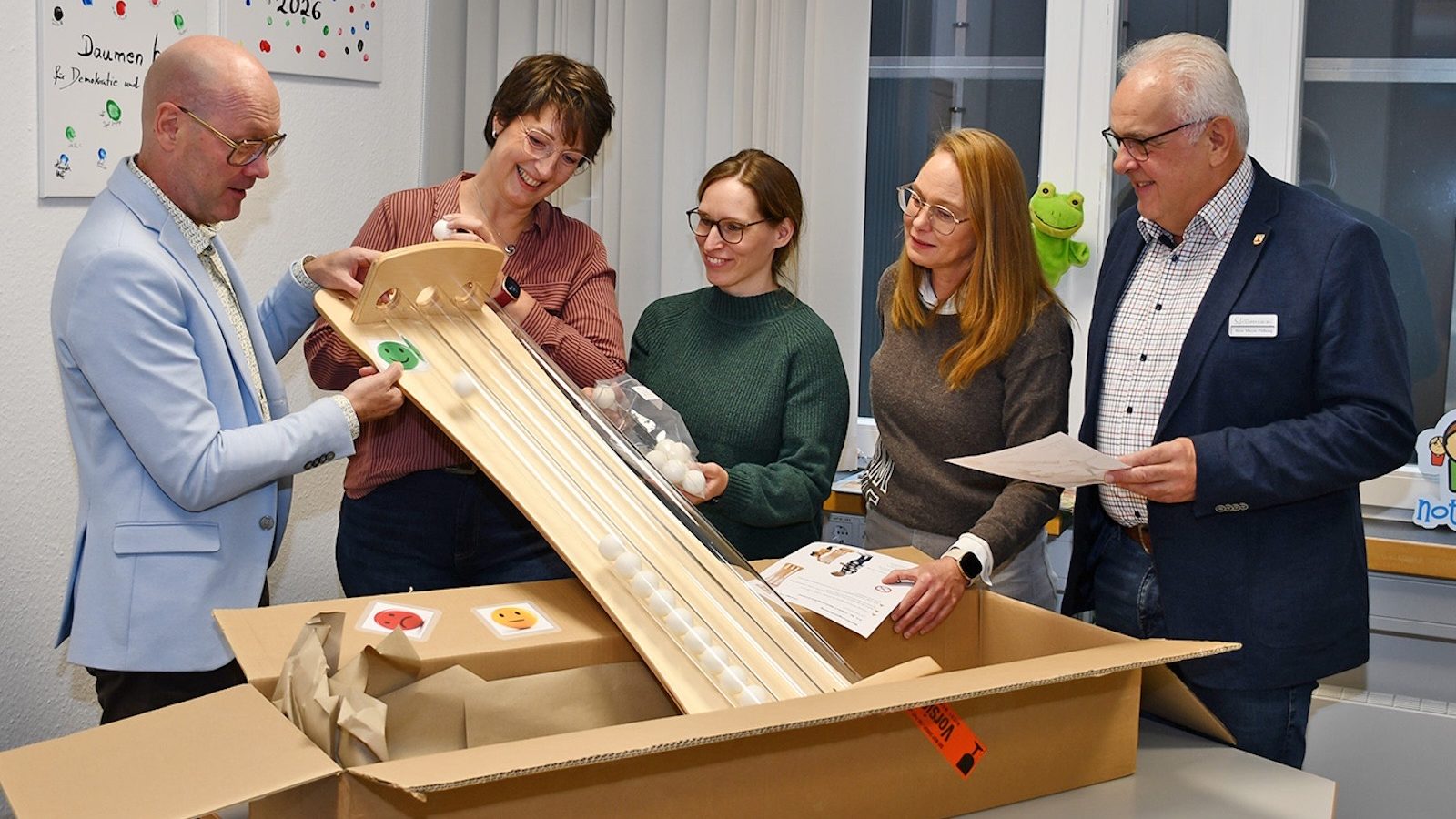 Fünf Personen packen eine hölzerne Abstimmungssäule mit Röhren aus einem Karton aus