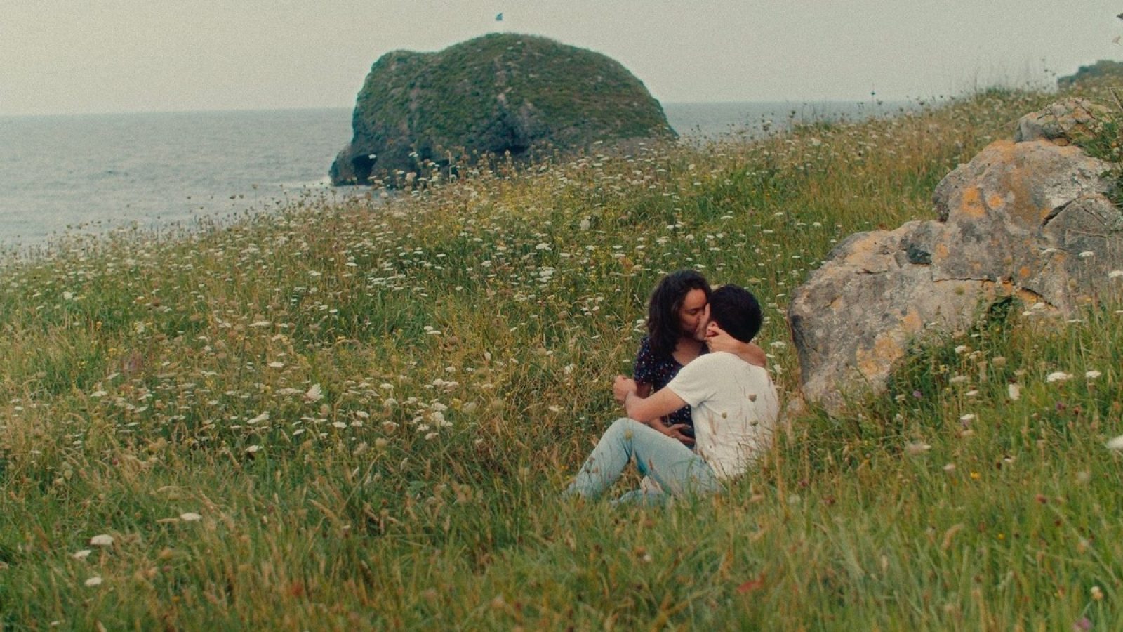 Zwei Personen sitzen im Gras am Meer und teilen einen intimen Moment in einer natürlichen Umgebung.