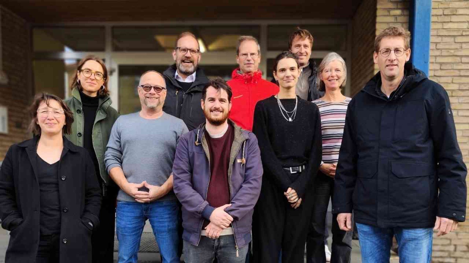 Gruppe von Personen, die vor einem Gebäude stehen, mit verschiedenen Gesichtsausdrücken und Kleidung.