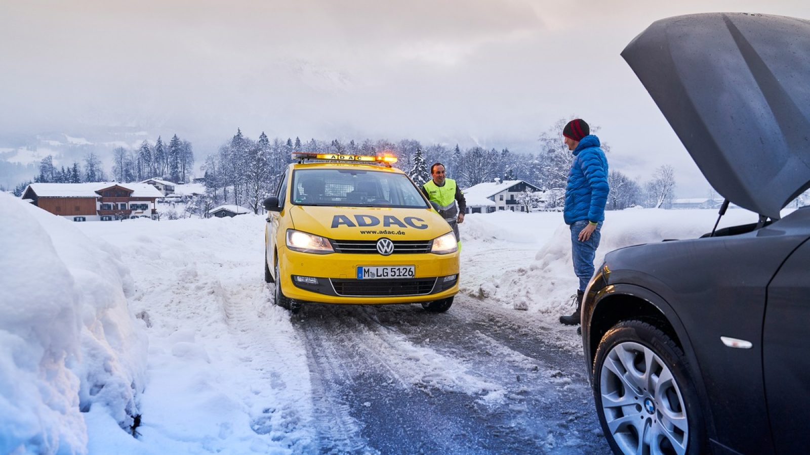 Gelbes ADAC-Fahrzeug mit Warnlichtern auf einer verschneiten Straße in den Alpen, während ein Mann mit einem anderen Auto spricht.