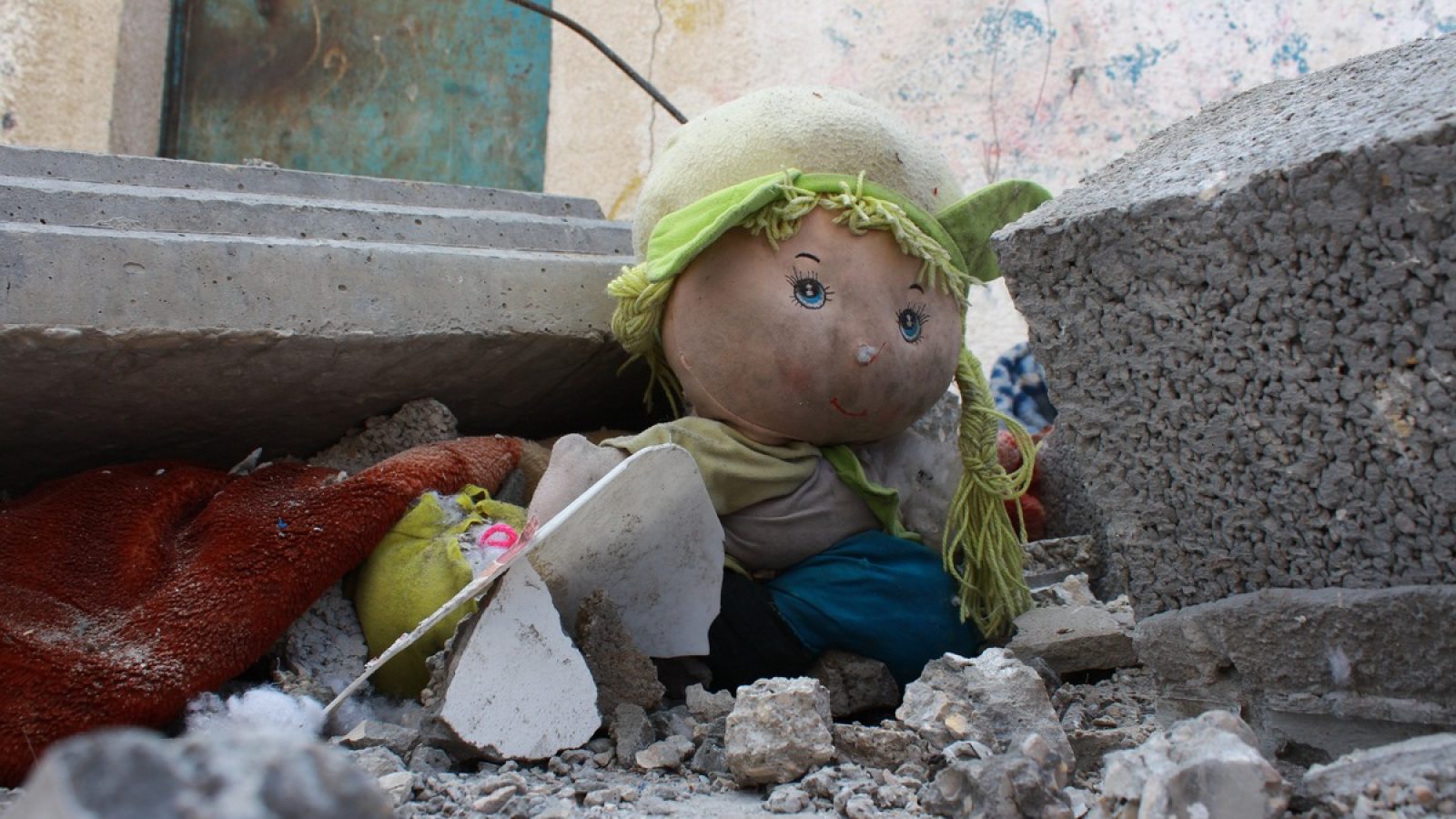 Eine beschädigte Puppe mit grüner Mütze und blonden Haaren, umgeben von Trümmern und Schutt.