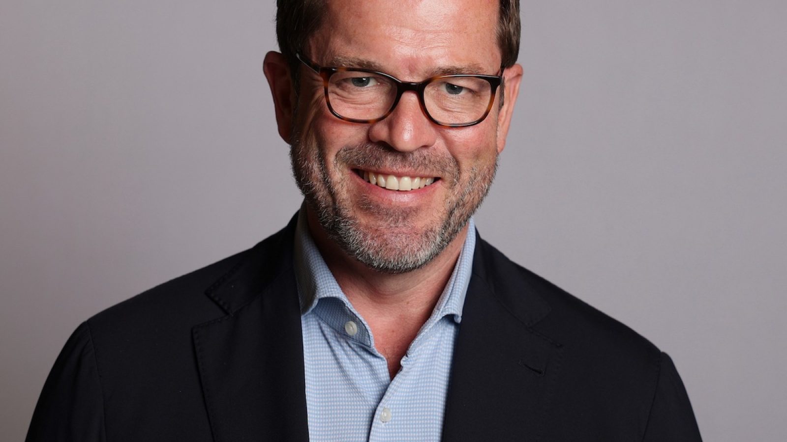 Mann mit Brille und Bart, der in einem dunklen Blazer und einem hellen Hemd lächelt.