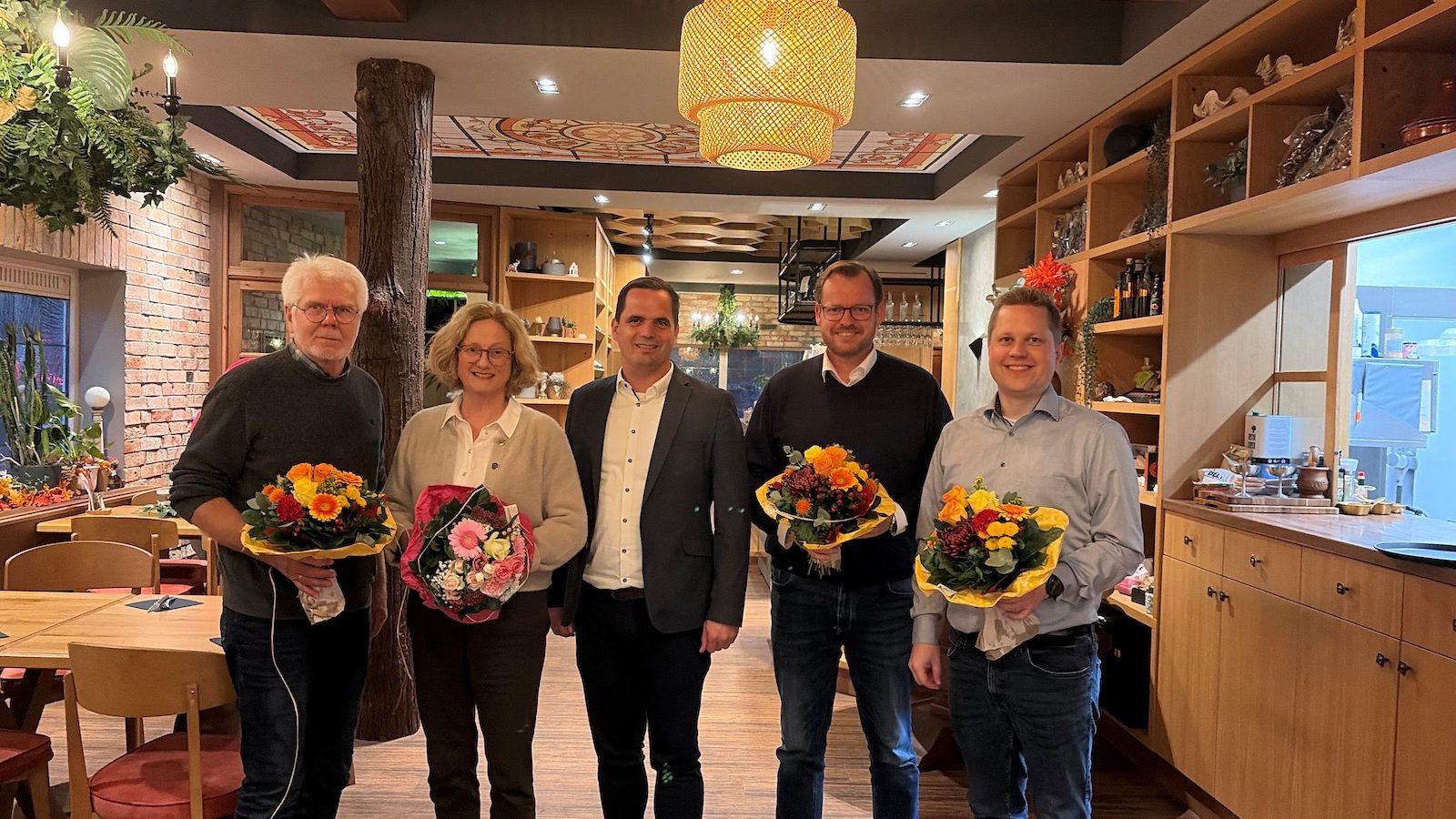 Fünf Personen stehen in einem Restaurant, vier von ihnen halten Blumensträuße