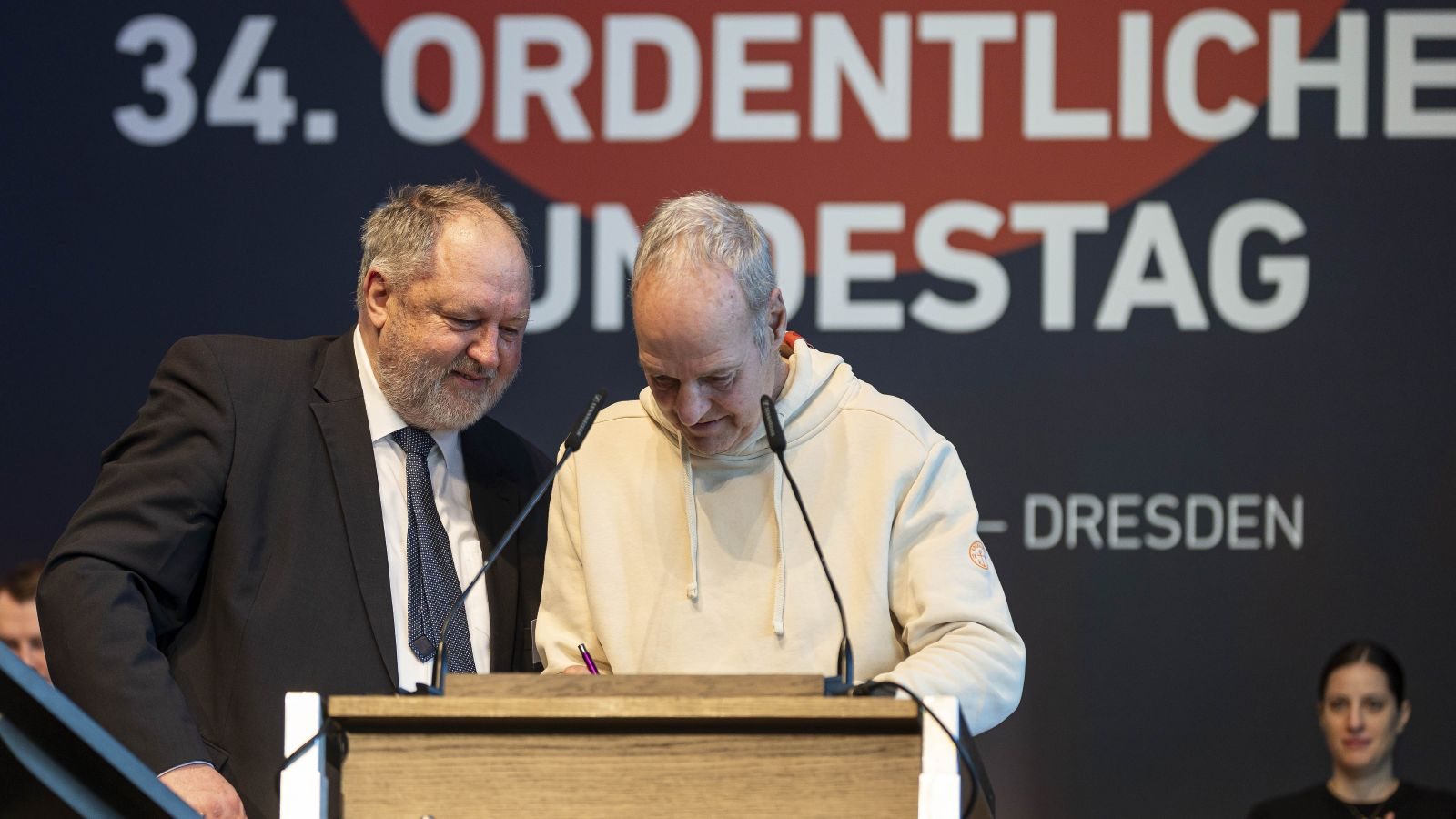 Zwei Männer stehen an einem Rednerpult während einer Veranstaltung in Dresden, einer schreibt etwas auf ein Blatt Papier.