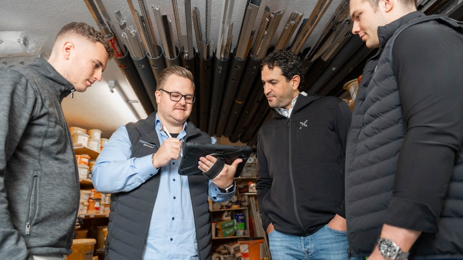 Vier Männer in einem Lagerraum, einer nutzt ein Tablet, während die anderen zuschauen und diskutieren.