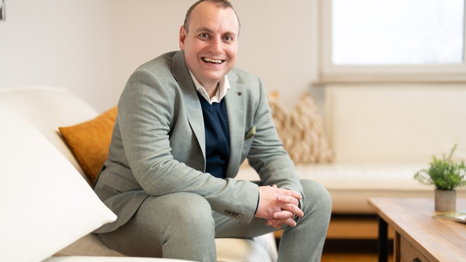 Mann in einem grauen Anzug sitzt auf einem Sofa und lächelt freundlich in die Kamera.