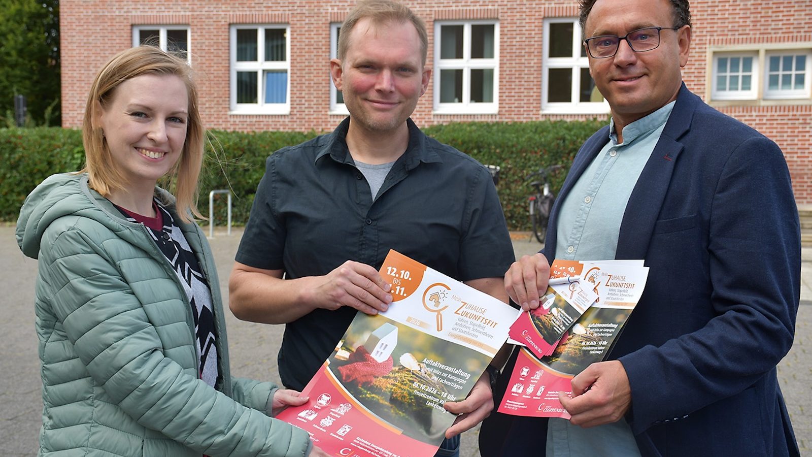 Vanessa Witt, Stefan Nienaber und Sergej Brandt halten Plakate der Kampagne Mein Zuhause zukunftsfit vor einem Backsteingebäude