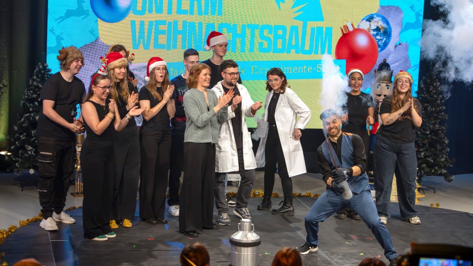 Gruppe von Menschen in Weihnachtsmützen, die auf einer Bühne mit einem Wissenschaftler und einem Experiment stehen.