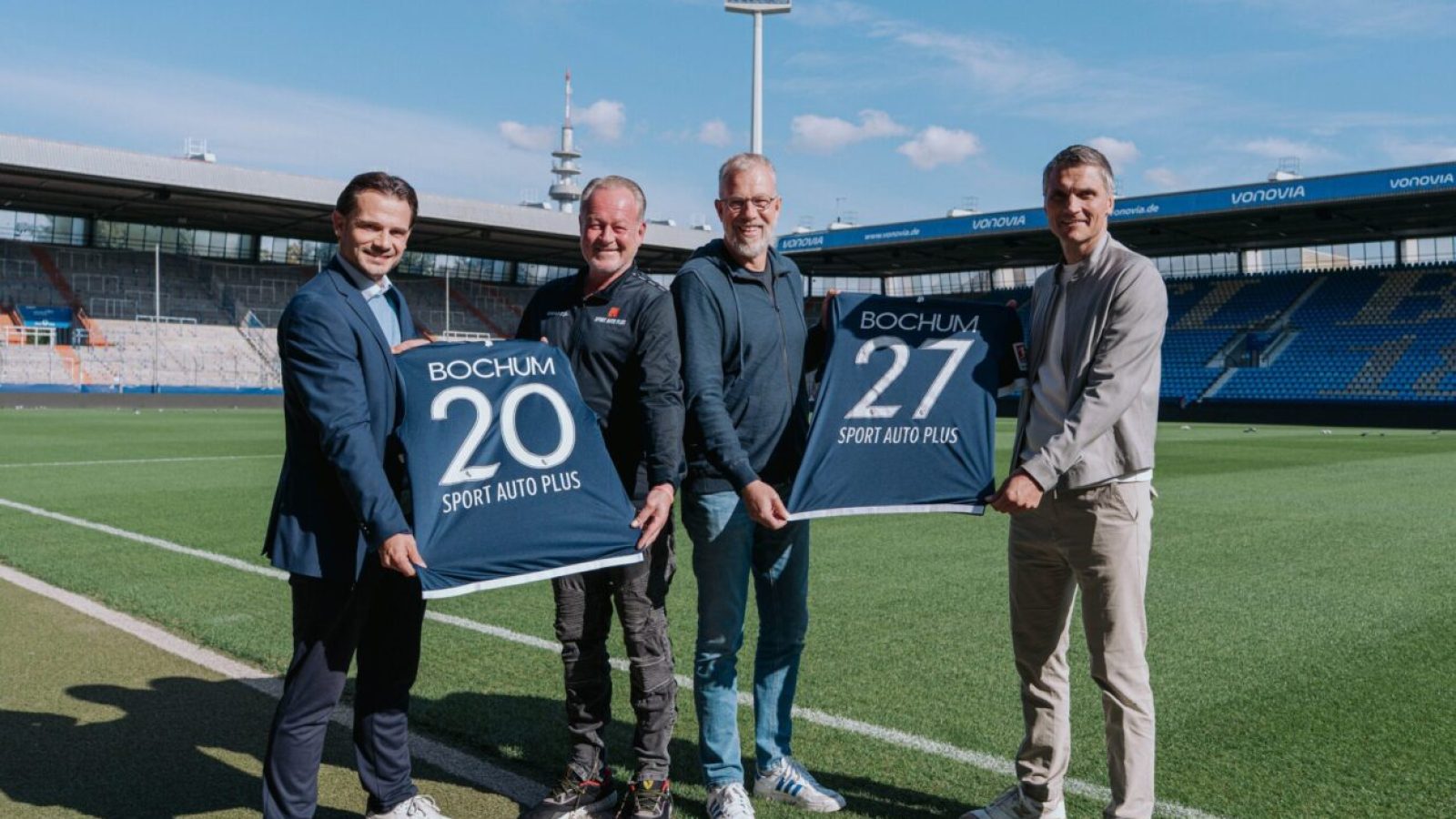Vier Männer posieren im Stadion mit Bochum-Trikots, Nummern 20 und 27, unter blauem Himmel. Sport Auto Plus Sponsoring.