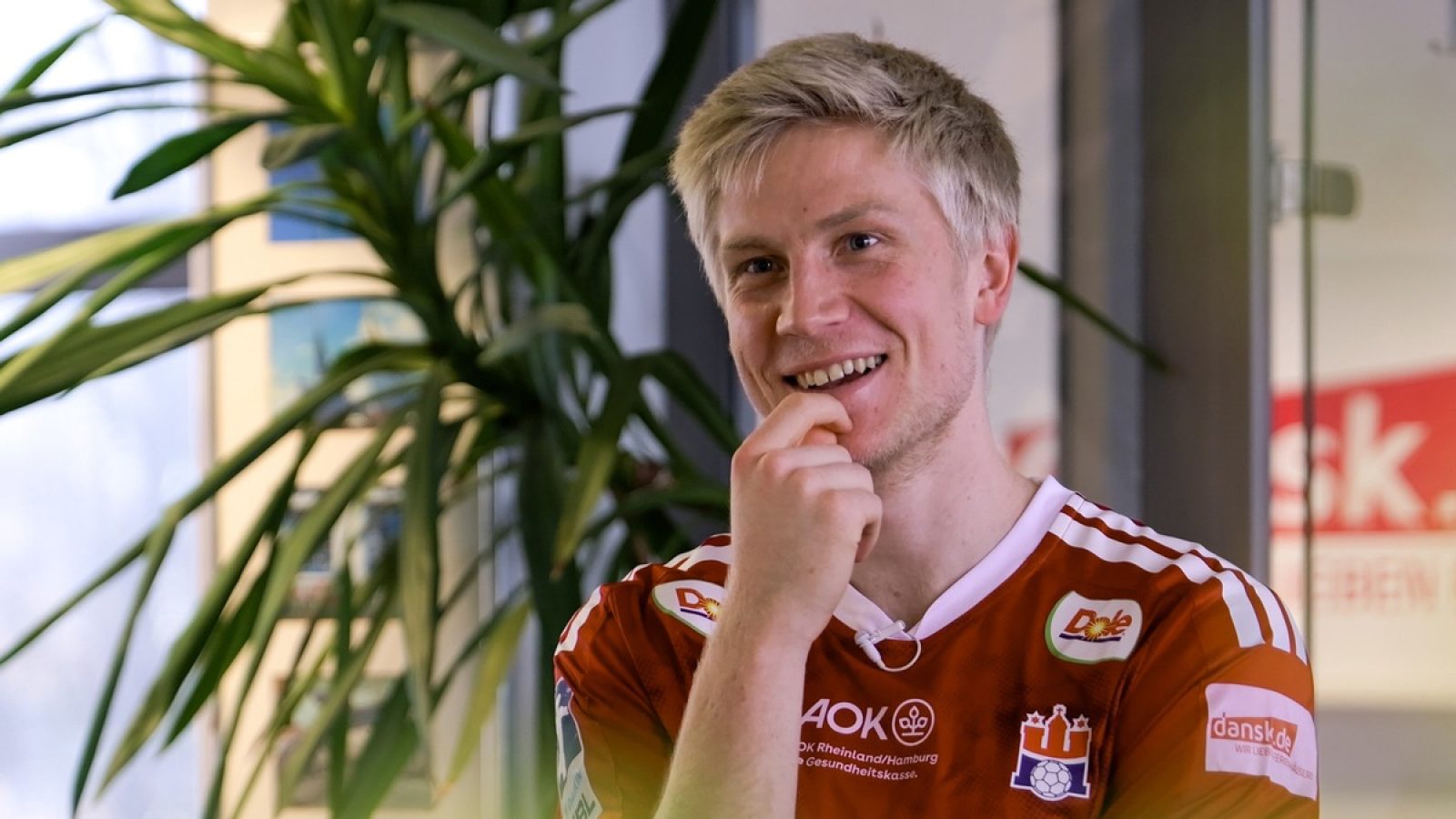 Lächelnder Mann mit kurzem, blonden Haar in einem rot-weißen Sporttrikot mit Sponsorenlogos.