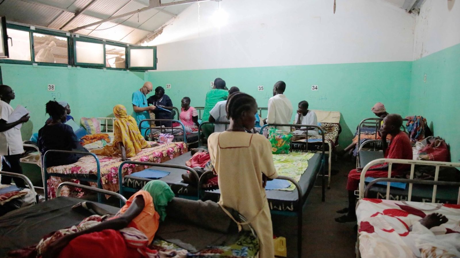 Patienten in einem Krankenhauszimmer mit mehreren Betten und medizinischem Personal, das sich um die Patienten kümmert.