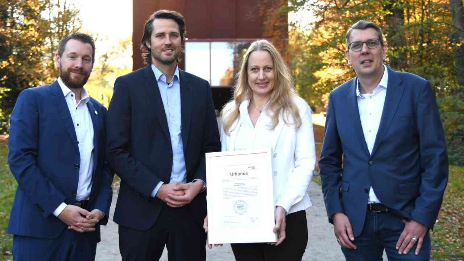 Vier Personen stehen im Freien und halten eine Urkunde in den Händen, umgeben von Bäumen und herbstlichem Laub.