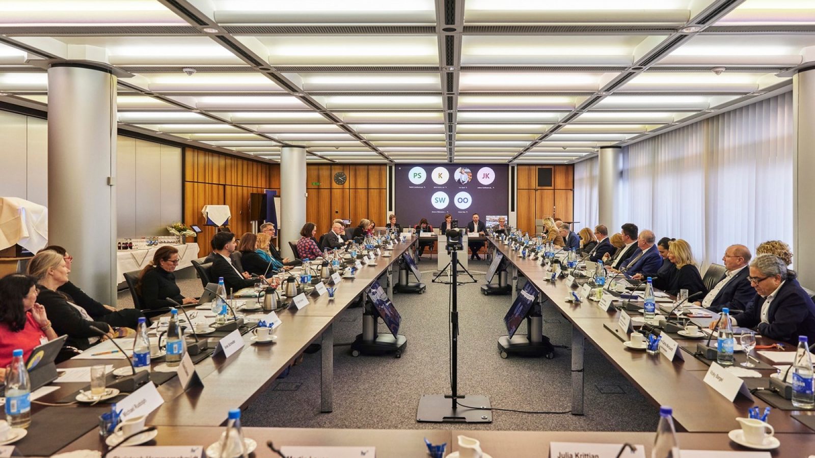 Konferenzraum mit einem langen Tisch, umgeben von Teilnehmern und einem Bildschirm im Hintergrund.