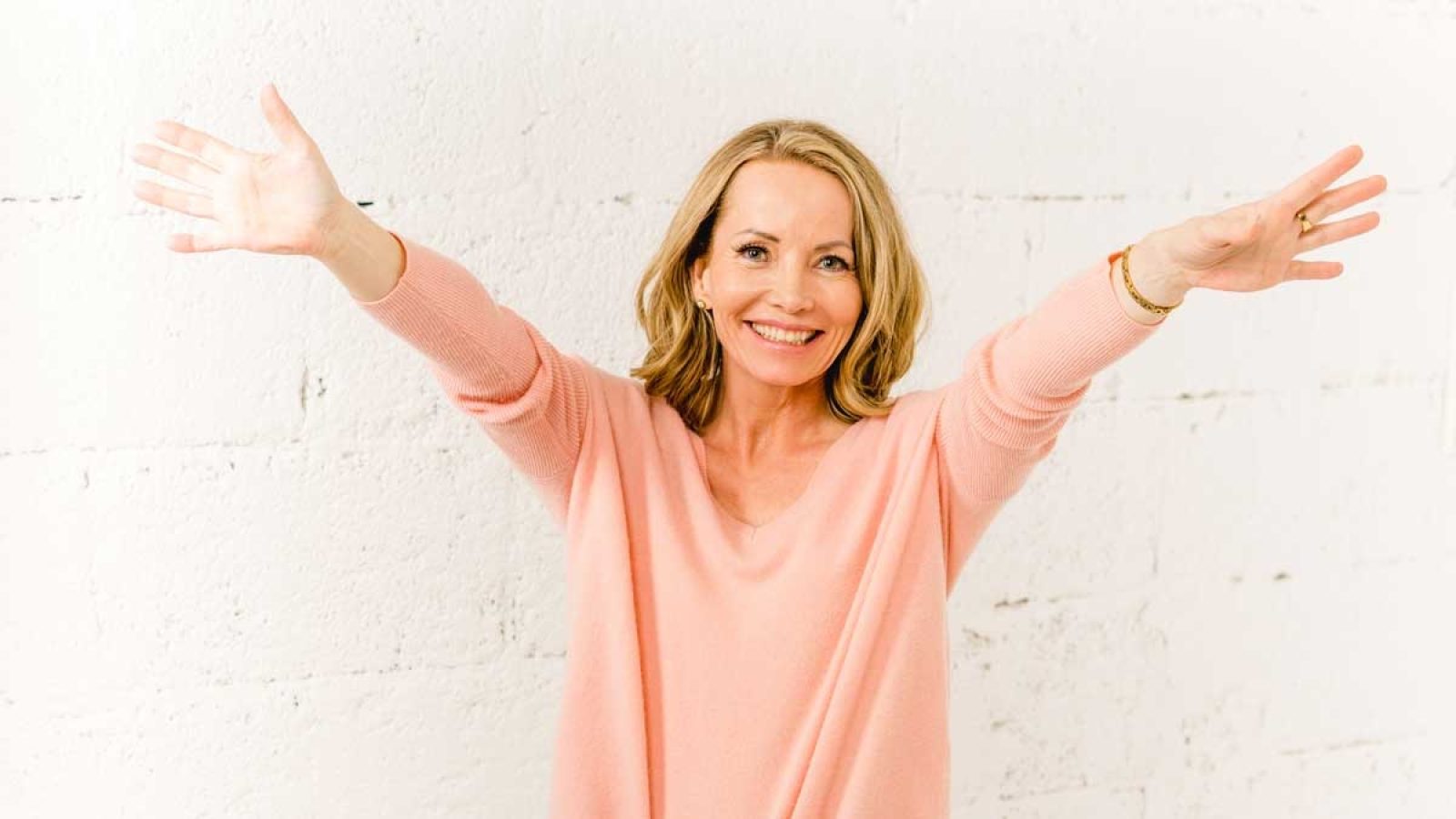 Frau mit blonden Haaren in einem rosa Pullover, die mit ausgebreiteten Armen lächelt.