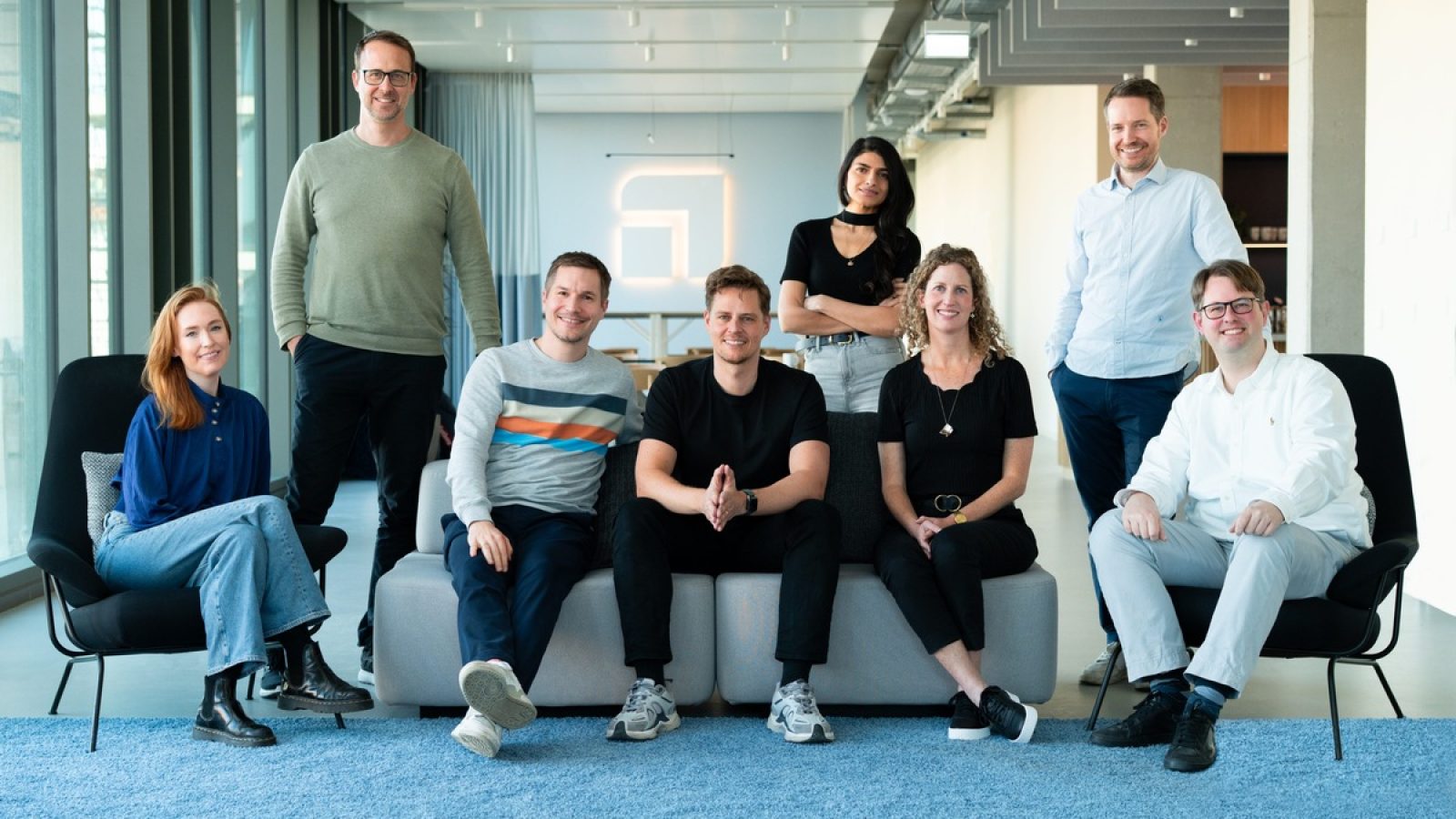 Eine Gruppe von acht Personen sitzt auf einem Sofa in einem modernen Büro mit großen Fenstern und einem blauen Teppich.