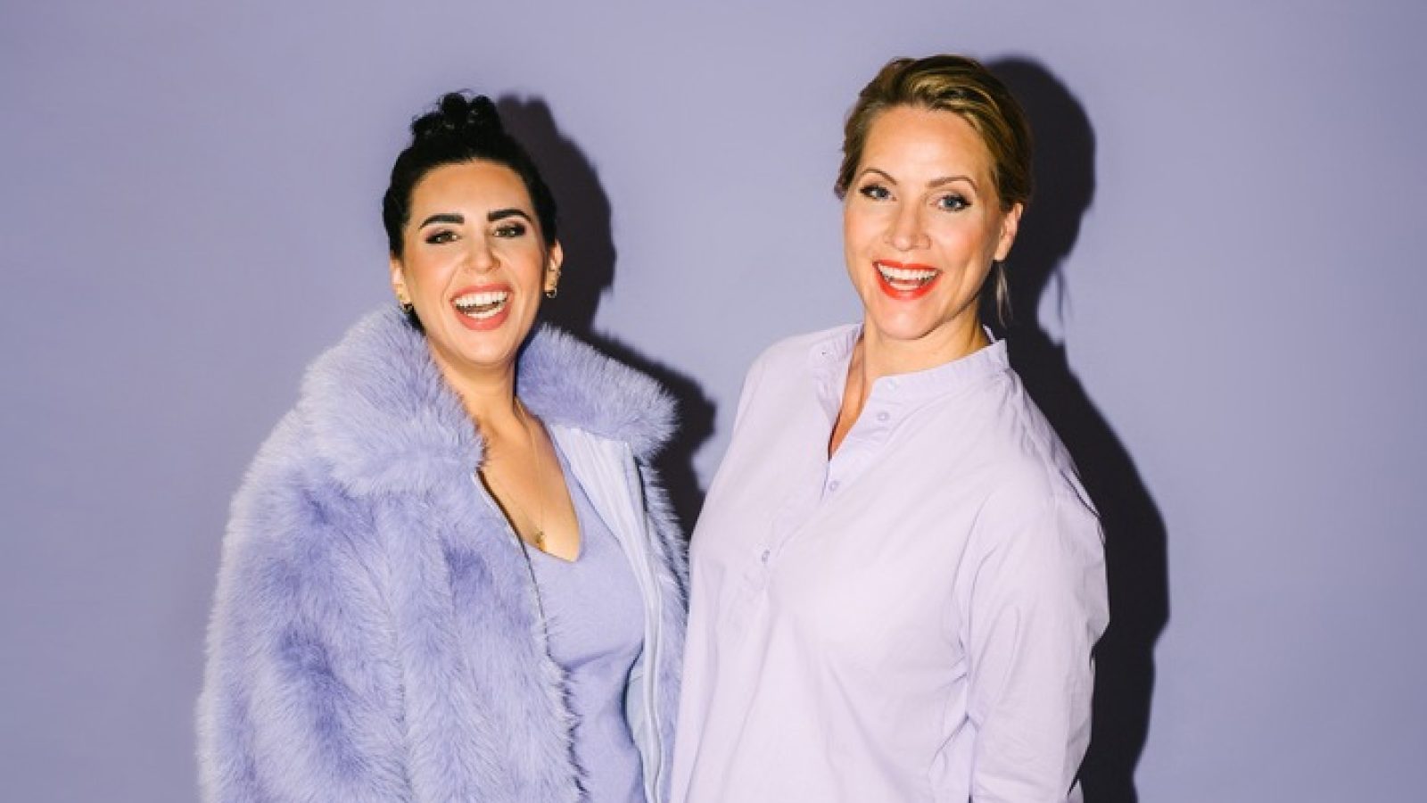 Zwei Frauen in lila Kleidung, eine trägt einen flauschigen Mantel, beide lächeln vor einem lila Hintergrund.