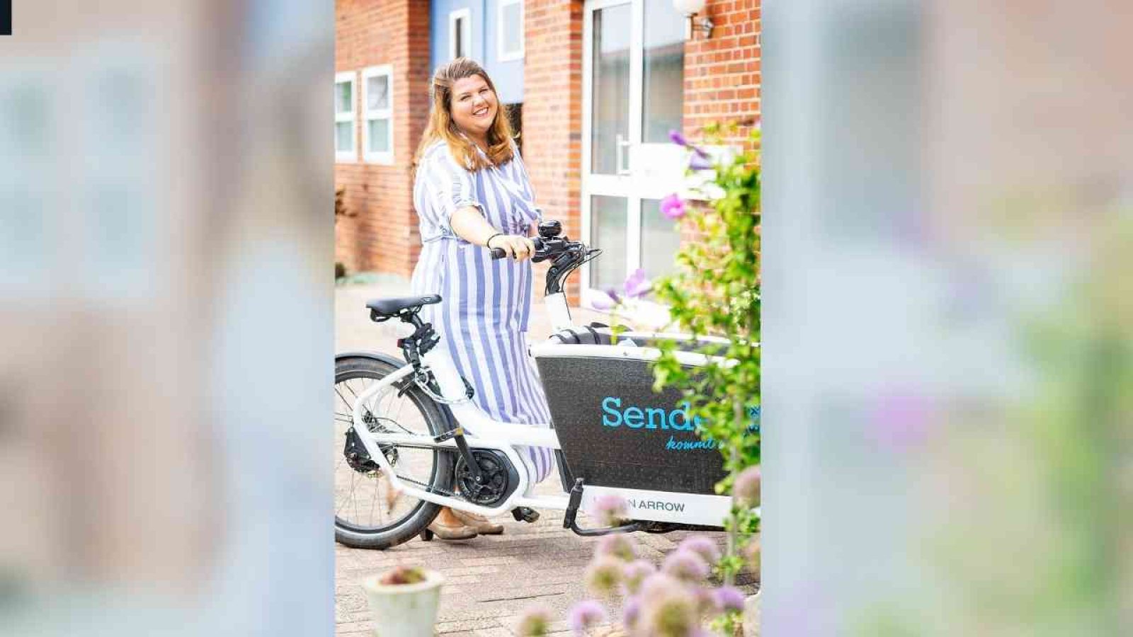 Frau mit gestreiftem Kleid steht neben einem Lastenrad mit der Aufschrift 'Senden'.