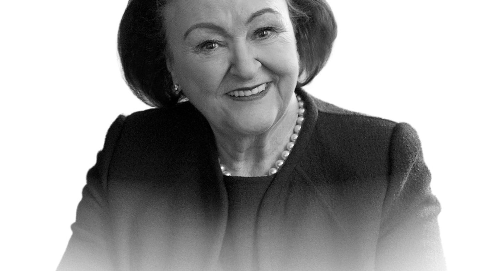 Porträt einer lächelnden Frau mit Perlenkette und dunkler Jacke, in einem neutralen Schwarz-Weiß-Stil.
