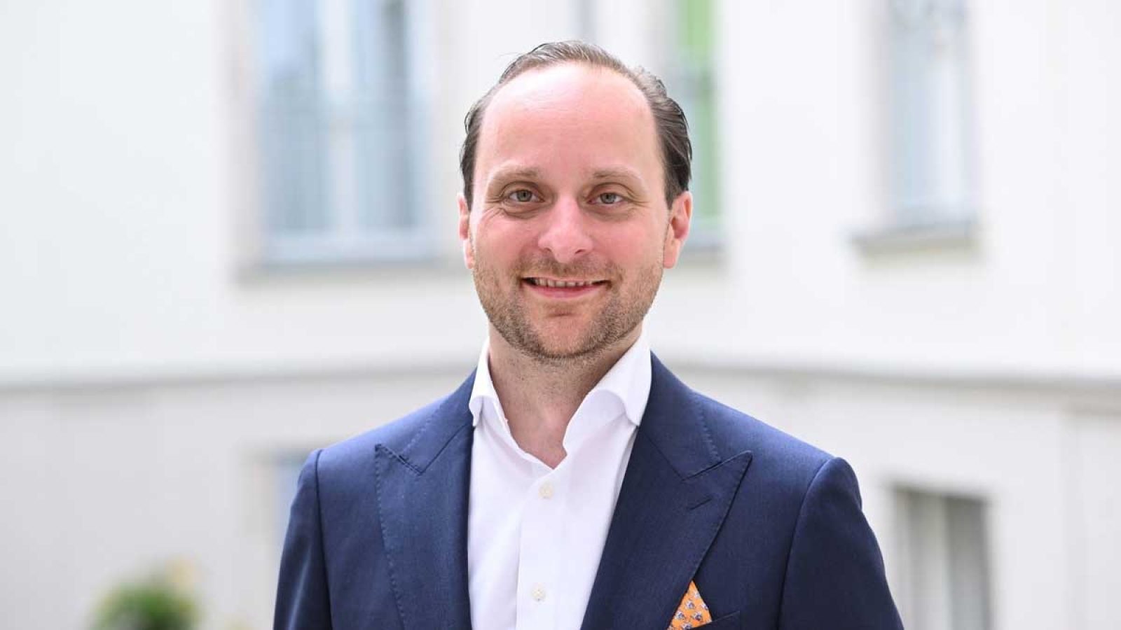 Mann in einem blauen Anzug mit einem weißen Hemd und einem orangefarbenen Einstecktuch, lächelt in die Kamera.