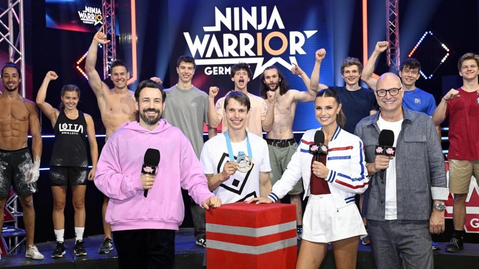 Gruppe von Teilnehmern und Moderatoren bei Ninja Warrior Germany, die auf einer Bühne posieren und jubeln.