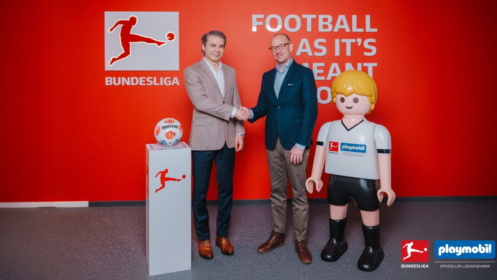Zwei Männer schütteln sich die Hände vor einer roten Wand mit dem Bundesliga-Logo und einem Playmobil-Figur in Fußballkleidung.