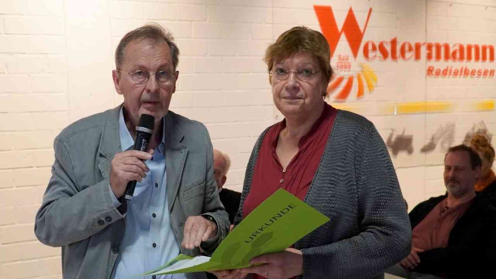 Zwei Personen bei einer Mitgliederversammlung, eine mit Mikrofon und die andere mit einer Urkunde in der Hand.