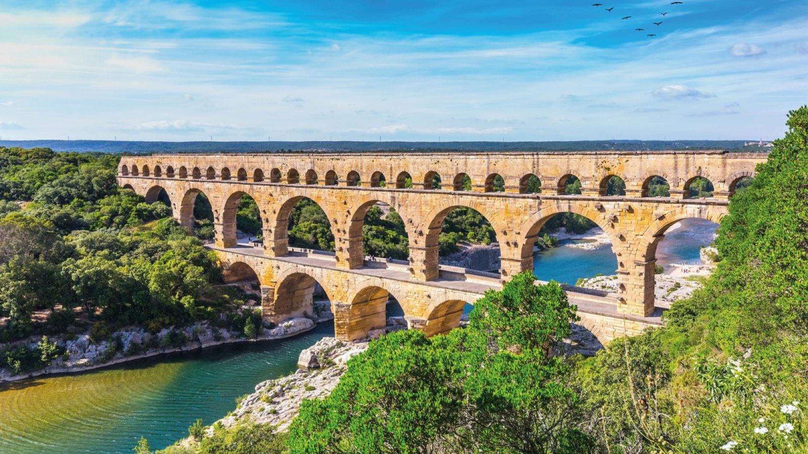 Römisches Aquädukt Pont du Gard mit mehreren Bögen über einem Fluss in einer grünen Umgebung.