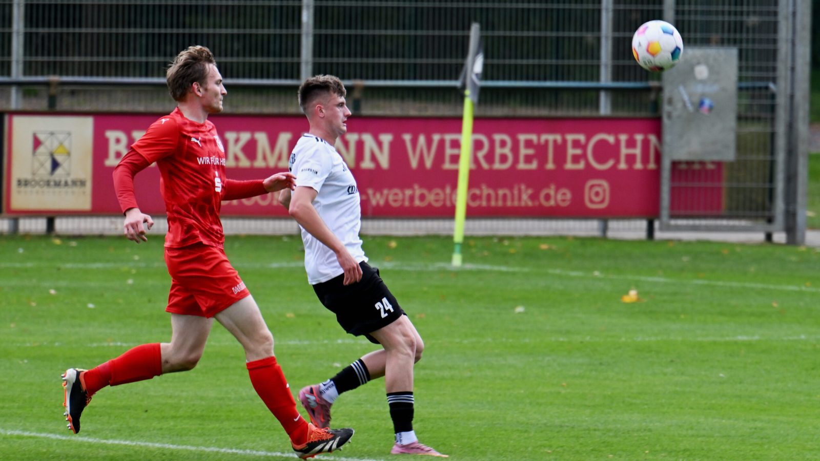 Zwei Fußballspieler im Spiel, einer in rotem Trikot, der andere in weißem, laufen zum Ball auf grünem Feld.