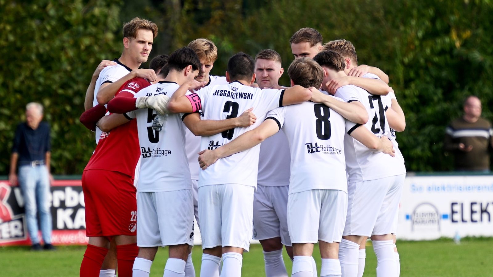 Spieler des SC Spelle-Venhaus in weißen Trikots stehen beim Derby in Hemsen im Kreis zusammen
