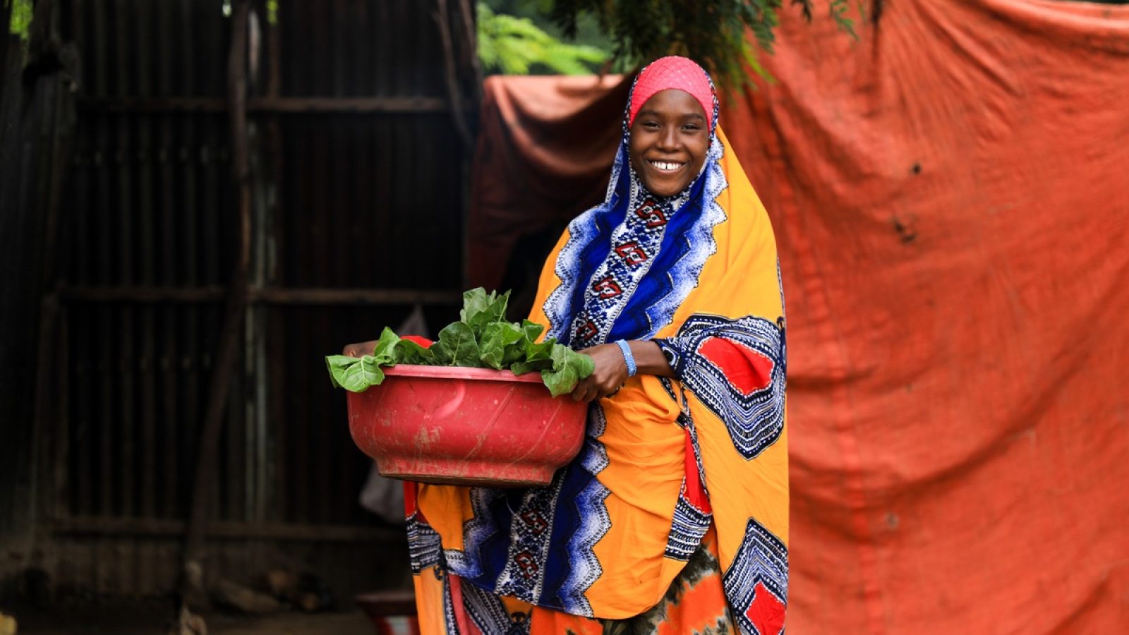 Frau in traditioneller Kleidung hält einen roten Behälter mit frischem Gemüse in der Hand.
