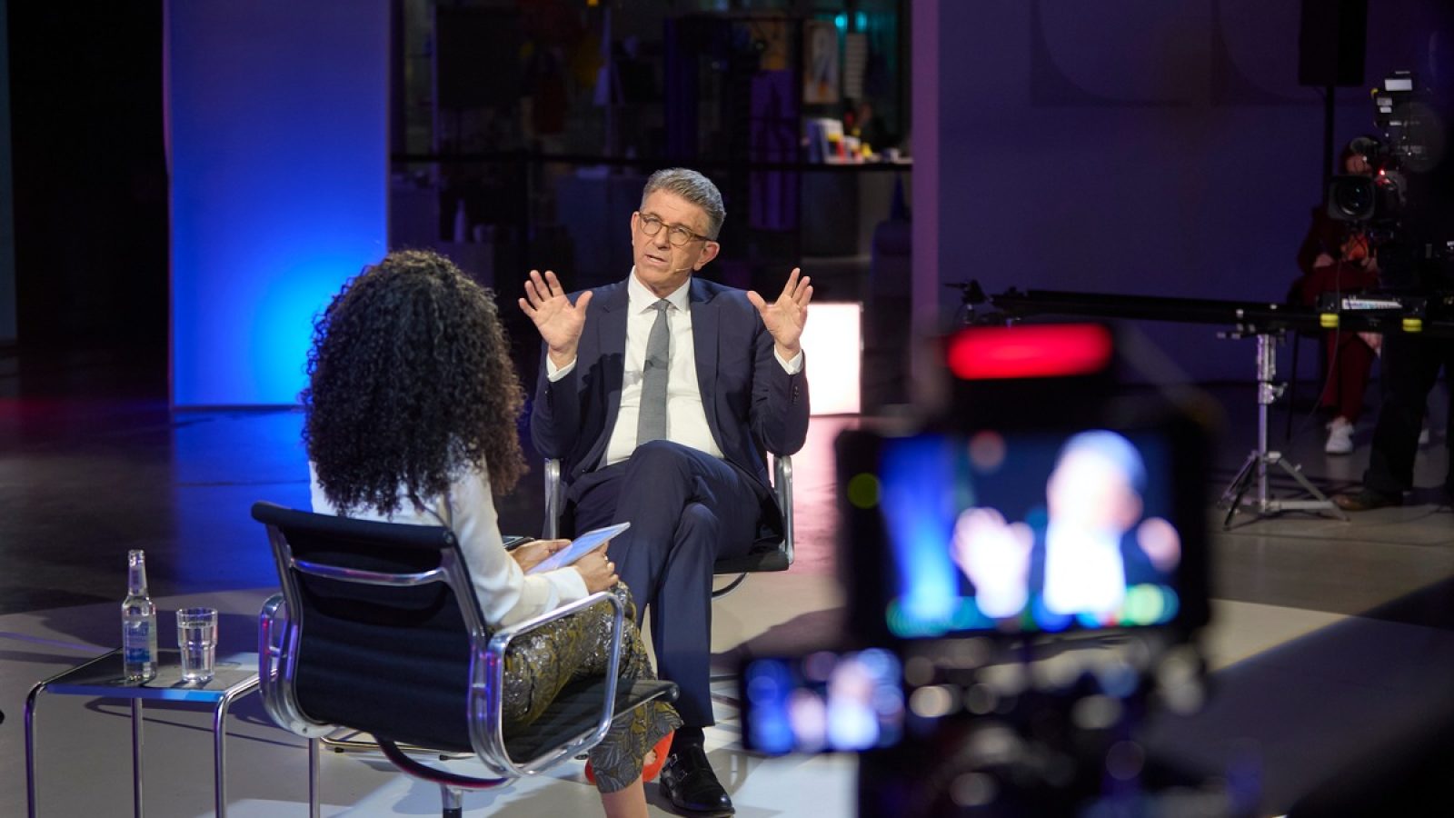 Mann in Anzug sitzt im Interview mit einer Frau, während eine Kamera auf sie gerichtet ist.