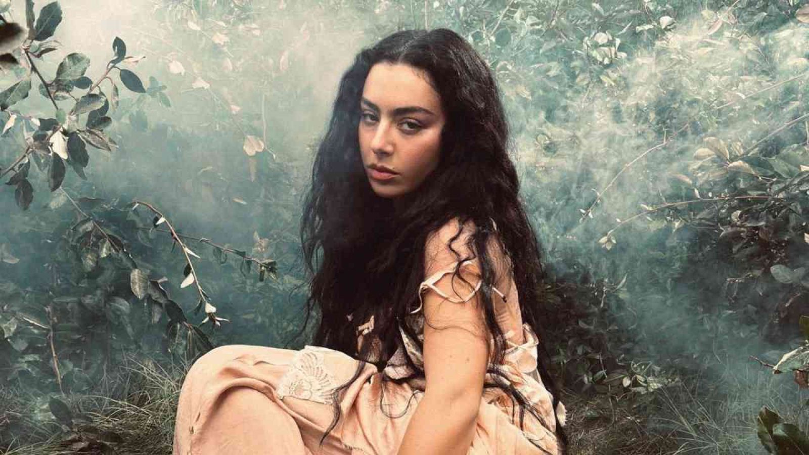 Frau mit langen, dunklen Haaren in einem fließenden Kleid, umgeben von Pflanzen und Nebel.