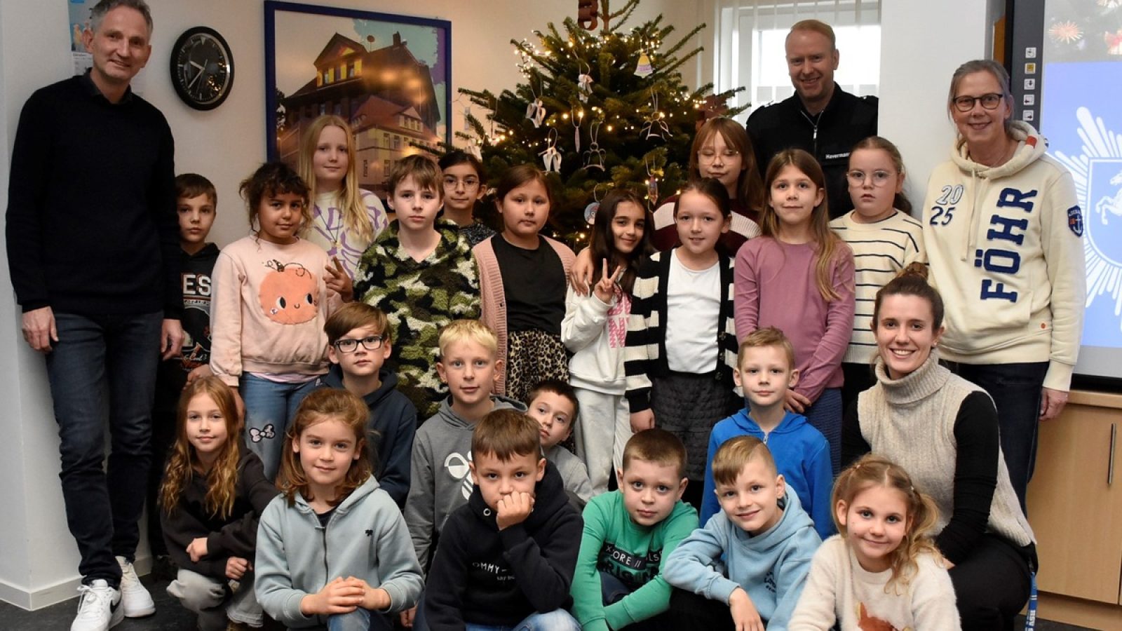 Gruppe von Kindern und Erwachsenen vor einem Weihnachtsbaum in einem Raum, umgeben von Dekorationen und festlicher Atmosphäre.