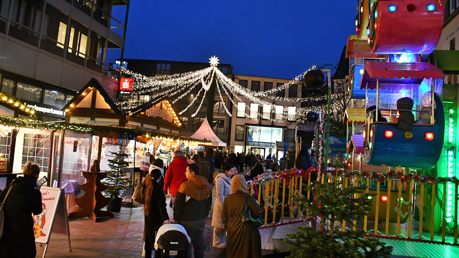 Weihnachtsmarkt mit festlich beleuchteten Ständen und Fahrgeschäften in einer Stadtstraße bei Dämmerung.