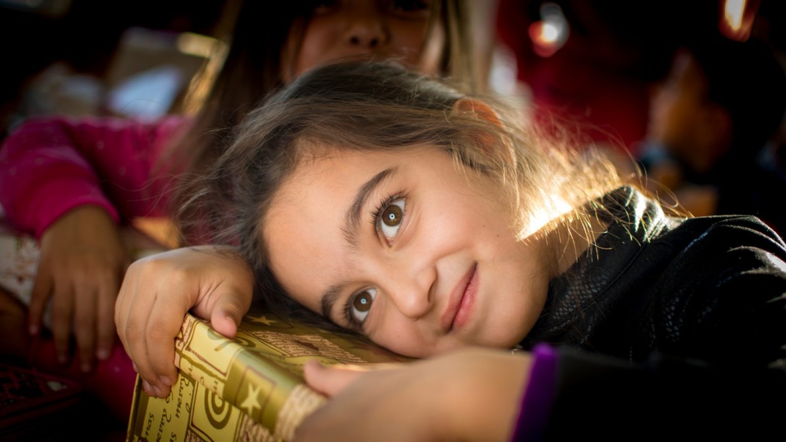 Ein Mädchen mit braunen Augen umarmt ein Geschenk mit goldener Verpackung und lächelt in die Kamera.