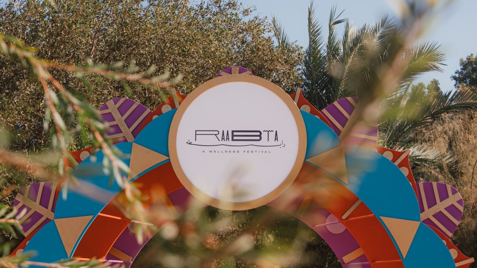 Bunte Bogenstruktur mit dem Logo des Rabta Wellness Festivals, umgeben von Pflanzen und Bäumen.