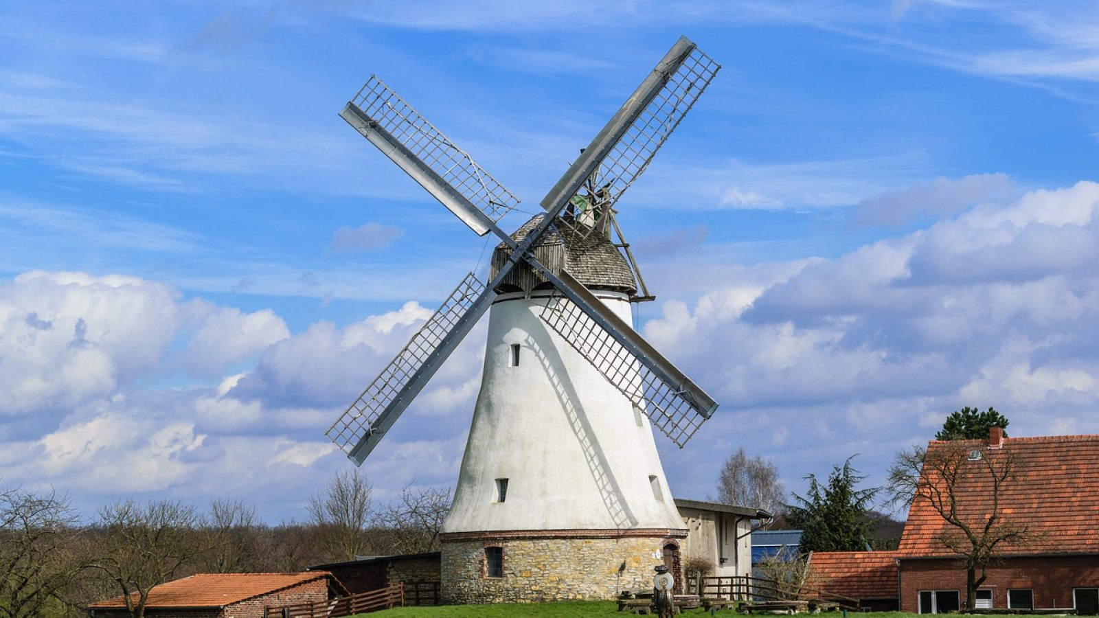 Windmühle mit weißen Flügeln und einem traditionellen Steingebäude im Hintergrund unter einem blauen Himmel.