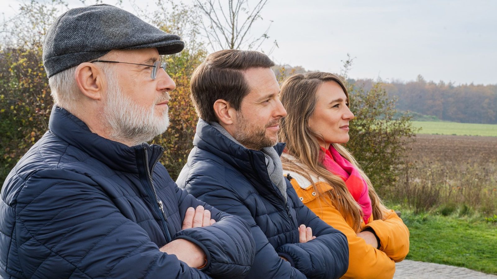 Drei Personen in Winterjacken stehen nebeneinander, mit verschränkten Armen, im Freien bei bewölktem Himmel.