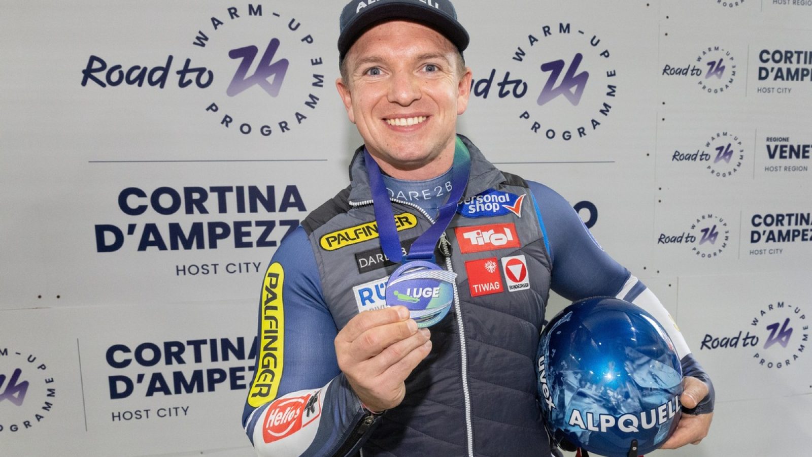 Wolfgang Kindl hält eine Medaille und einen Helm in der Hand, während er vor einem Hintergrund mit dem Logo von Cortina d'Ampezzo steht.